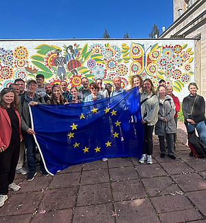 Eine Gruppe von etwa 20 Personen steht im Freien vor einer farbenfrohen Wandmalerei mit folkloristischen Motiven. In der Mitte halten einige Personen eine Europaflagge mit gelben Sternen auf blauem Grund. Es ist ein sonniger Tag, die Menschen wirken fröhlich und posieren für das Gruppenfoto.