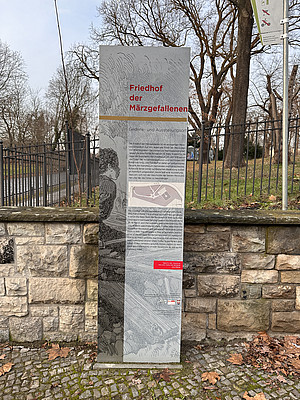 Eine Infotafel aus Metall mit der Aufschrift „Friedhof der Märzgefallenen“ steht vor einer niedrigen Steinmauer und einem Zaun. Die Tafel enthält Informationen zur historischen Gedenkstätte in Textform sowie eine Karte. Im Hintergrund sind Bäume ohne Laub zu sehen.