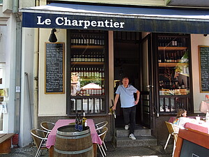 Ein bistroartiges Lokal mit zwei schmalen Fenstern mit vielen Weinflaschen und einer schmalen Tür, in der ein Mann steht. Davor Esstische mit rot-weiß karierten Tischdecken, die von einer blauen Markise mit der Aufschrift "Le Charpentier" beschattet werden. 
