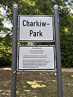 Ein zweigeteiltes Schild steht auf einer Wiese vor grünem Baumbestand. Oben steht in großen schwarzen Buchstaben: „Charkiw-Park“. Darunter befindet sich eine Texttafel mit Informationen auf Deutsch: Sie erklärt, dass Charkiw eine Stadt in der Ukraine ist, seit dem 24.10.1990 Partnerstadt des Berliner Bezirks Steglitz-Zehlendorf, und erinnert an die Opfer des russischen Angriffskrieges ab dem 24. Februar 2022. Besonders hervorgehoben wird Boris Romantschenko, ein Holocaust-Überlebender und langjähriger Gedenkarbeiter. An den Pfosten des Schilds sind blau-gelbe Bänder als Zeichen der Solidarität mit der Ukraine befestigt.