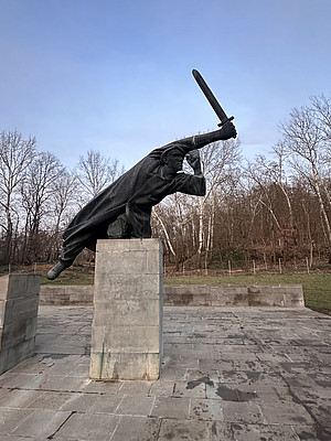Eine monumentale Bronzestatue zeigt einen kämpfenden Mann mit erhobenem Schwert, der nach vorne stürmt. Die Figur steht auf einem hohen rechteckigen Sockel, im Hintergrund sind kahle Bäume und blauer Himmel zu sehen. Das Denkmal erinnert an die Interbrigadisten.