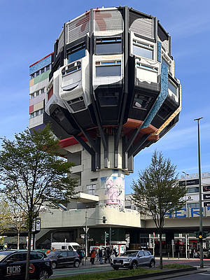 Ein futuristisch anmutendes, farbenfrohes Gebäude mit achteckiger Struktur thront auf einer zentralen Betonsäule über einer belebten Straßenkreuzung. Das Gebäude, bekannt als der „Bierpinsel“ in Berlin, weist eine einzigartige Architektur mit vorgelagerten Fenstern und bunten Fassadenelementen auf. Unten fahren Autos und Fußgänger überqueren die Straße. Im Hintergrund sind moderne Geschäftsgebäude und Schilder erkennbar. Ein blauer Himmel und grüne Bäume rahmen die Szene.