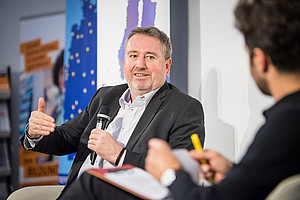 Ein Mann mittleren Alters mit grauen Haaren und Bart sitzt auf einem Podium, spricht in ein Mikrofon und gestikuliert mit der freien Hand. Er trägt ein weißes Hemd und ein dunkles Sakko. Ihm gegenüber sitzt eine weitere Person, die nur von hinten zu sehen ist und einen gelben Stift sowie ein Notizbuch hält. Im Hintergrund sind farbige Plakate mit europäischen Motiven sichtbar.